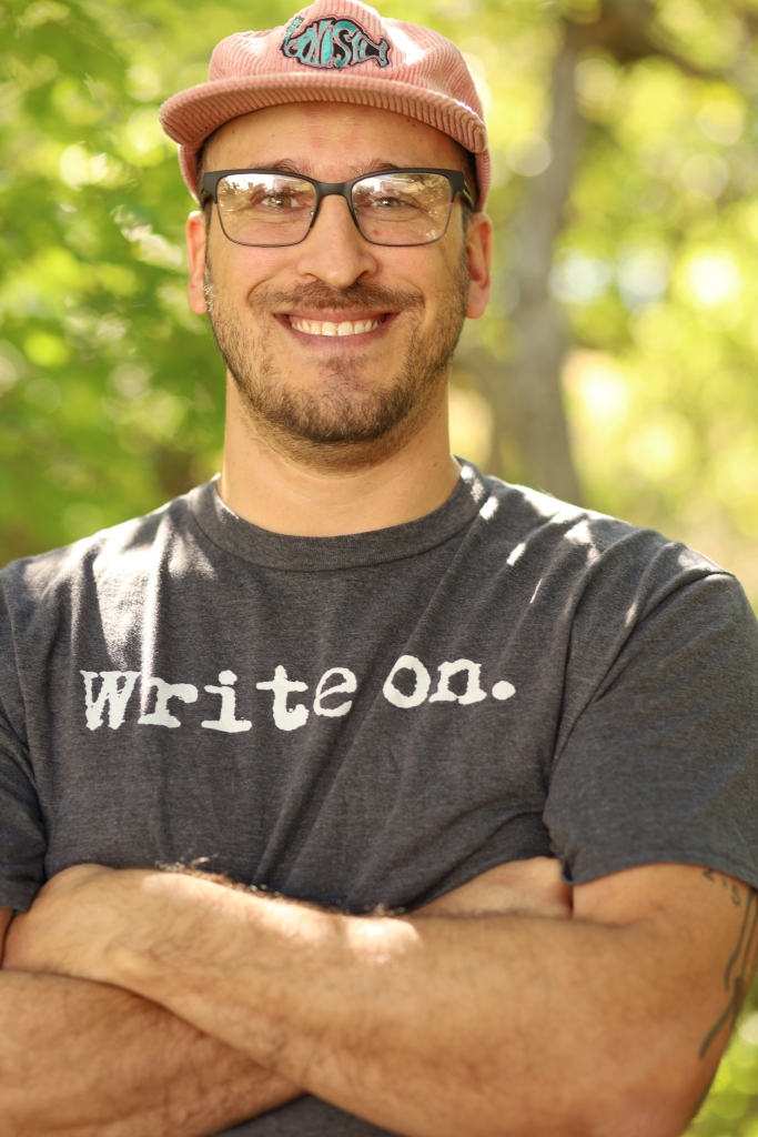 a photo of steve with arms crossed, a "write on" tee shirt, and glasses. He's smiling. He's wearing a pink corduroy hat with a "phish" band emblem on it.